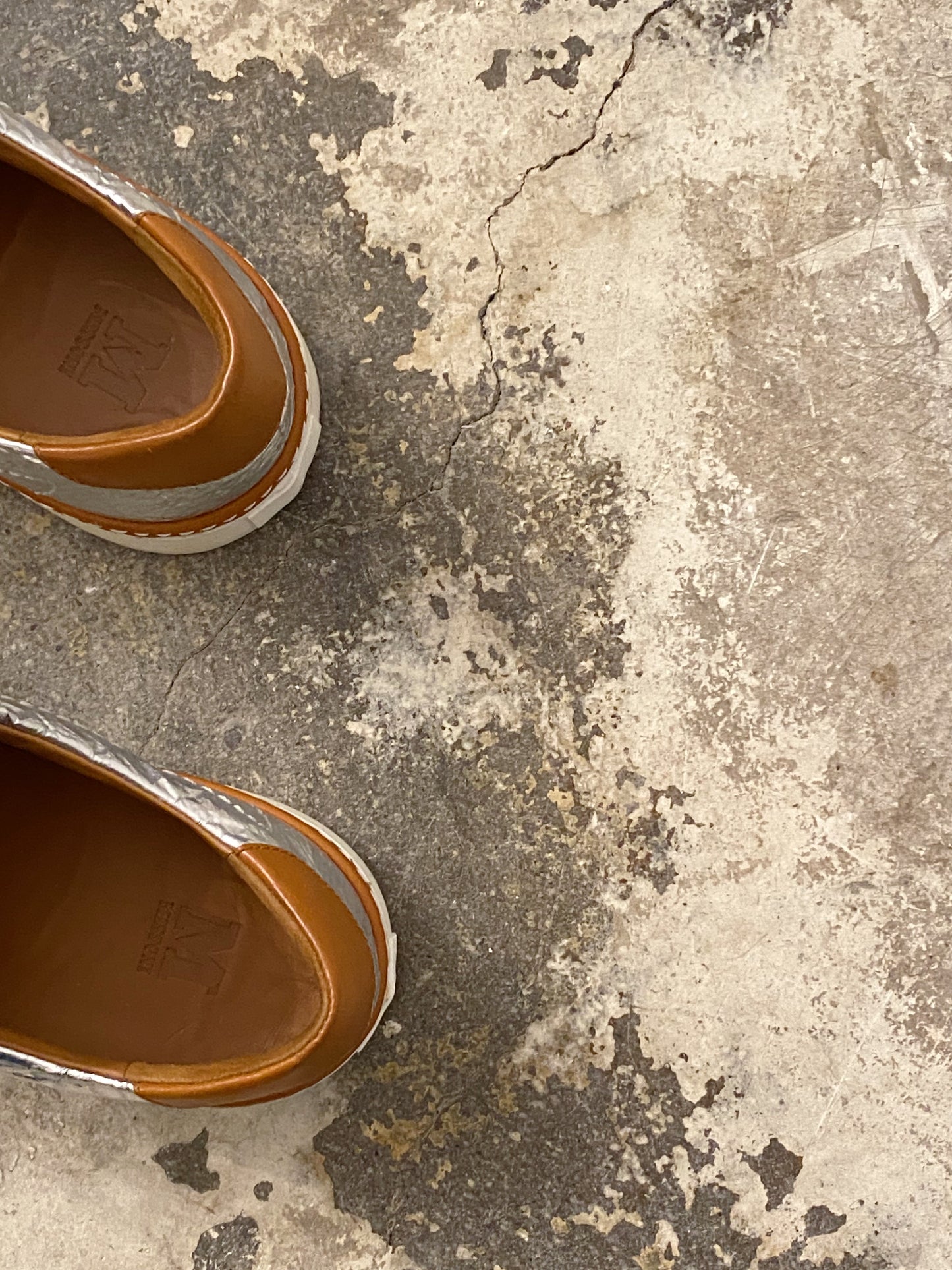 Close-up of a brown leather shoe on a textured concrete floor. M by Missoni silver & bronze lamé low top sneakers