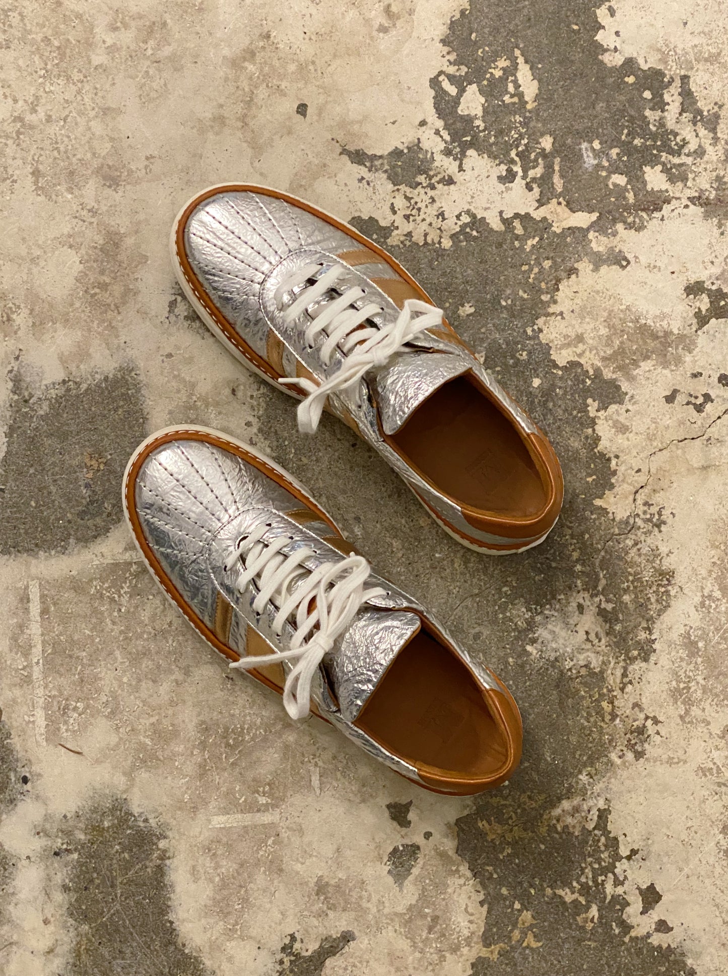 Pair of silver metallic shoes with brown soles on a textured surface. M by Missoni silver & bronze lamé low top sneakers